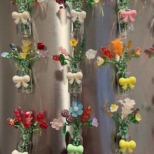 Op de afbeelding: Een verzameling miniatuur glazen vazen, elk gevuld met kleurrijke kralenbloemen. De vazen hebben witte strikaccenten. De bloemen zijn in verschillende kleuren, waaronder rood, roze, geel en blauw. De vazen zijn bevestigd aan een metalen oppervlak.