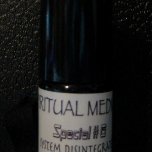 May include: A small, cylindrical bottle with a black cap and a white label. The label reads "RITUAL MED" above "Special #6" and "SYSTEM DISINTEGRAT". A symbol is printed at the bottom. The bottle is on a dark surface.