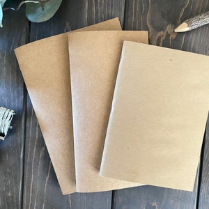 Könnte beinhalten: Drei braune Kraftpapier-Notizbücher, die auf einer Holzoberfläche gestapelt sind. Die Notizbücher sind leer und haben eine raue Textur.