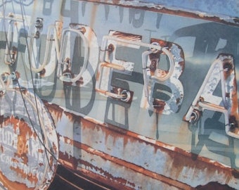 May include: A vintage, weathered sign with the word "SUNDERBA" in faded white letters with neon tubing. The sign is rusty and has a blue and gray background.