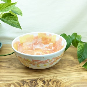May include: A white ceramic bowl with a pink and yellow watercolor design. The bowl is sitting on a wooden surface.
