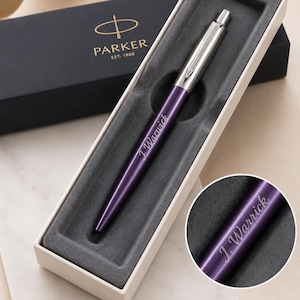 May include: A purple Parker ballpoint pen with a silver clip and cap, engraved with the name J. Warrick. The pen rests in a rectangular cream-colored box with a dark gray interior, accompanied by a black Parker gift box with gold text.
