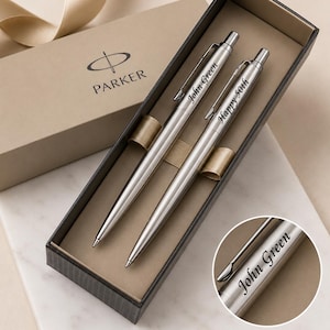 May include: A set of two silver-toned Parker ballpoint pens housed in a rectangular gift box. One pen is engraved with the name John Green, and the other is engraved with the text Happy 50th. The pens feature a metallic finish and a pocket clip.