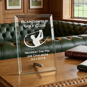 May include: A clear glass award with the text "BLANCHWOLD GOLF CLUB" and "NEAREST THE PIN JOE CHALMERS JULY 2018". The award sits on a wooden table with a silver mug, a book, and a golf towel. The room has a green leather sofa.