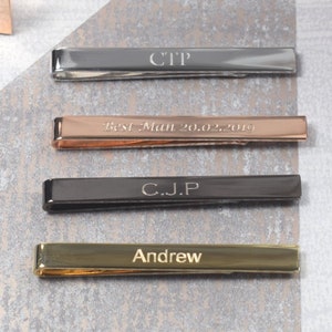 May include: Four tie bars in different colours and finishes. The top tie bar is silver with the initials "CTP" engraved on it. The second tie bar is rose gold with the text "Best Man 20.02.2019" engraved on it. The third tie bar is black with the initials "C.J.P" engraved on it. The bottom tie bar is gold with the name "Andrew" engraved on it.