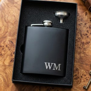 Puede incluir: Una petaca negra con las iniciales "WM" grabadas en la parte delantera, presentada en una caja negra con un pequeño embudo. La petaca es de metal y tiene una tapa plateada. La caja está sobre una superficie de madera.