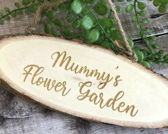 May include: A wooden sign with the text "Mummy's Flower Garden" engraved on it. The sign is oval shaped and has a rustic appearance.