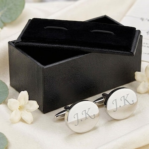 Może przedstawiać: Srebrne spinki do mankietów z inicjałami "J.K." wygrawerowanymi na każdej okrągłej powierzchni, prezentowane obok otwartego czarnego pudełka z aksamitnym wnętrzem. Białe kwiaty i zielone liście dopełniają scenę.