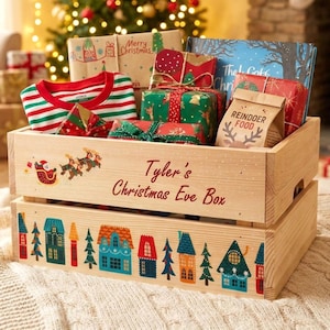 Könnte beinhalten: Eine helle Holzkiste mit der Aufschrift Tyler's Christmas Eve Box, einem Weihnachtsschlitten-Motiv und einer bunten Dorf-Illustration. Die Kiste ist gefüllt mit verpackten Geschenken, einem gestreiften Oberteil und einer Tüte mit der Aufschrift Reindeer Food.