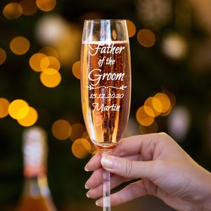 May include: A clear champagne flute with a personalized inscription that reads "Father of the Groom 15.12.2020 Martin". The flute is filled with sparkling wine and is being held by a hand.