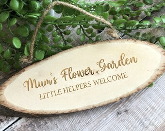 May include: A wooden sign with the text "Mum's Flower Garden Little Helpers Welcome" engraved in gold lettering. The sign is oval shaped and has a rustic, natural look.