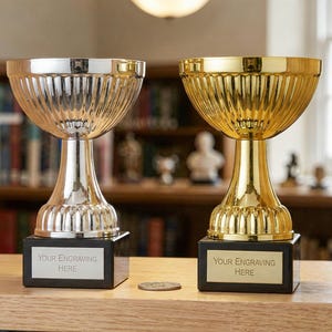 Puede incluir: Dos trofeos, uno plateado y otro dorado, están sobre una estantería de madera. Cada trofeo tiene un cuenco acanalado y una base de pedestal, montada sobre un cuadrado negro. El trofeo plateado tiene una base plateada, mientras que el dorado tiene una base dorada. Ambos tienen una etiqueta que dice "YOUR ENGRAVING HERE."