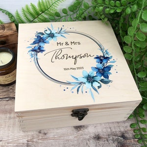 May include: A wooden box with a hinged lid and a latch closure. The lid is decorated with a blue and white floral wreath design and the text "Mr & Mrs Thompson 15th May 2023".
