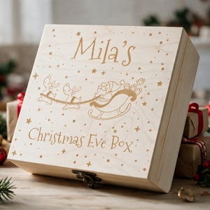 May include: A light-coloured wooden box with a hinged lid and a metal latch. The lid features an engraved design of Santa in a sleigh pulled by reindeer, surrounded by stars, with the text "Mila's Christmas Eve Box" centred on the front.