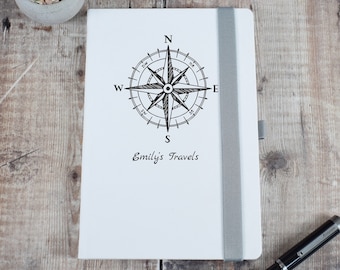 May include: White notebook with a black compass design and the text "Emily's Travels". A silver pen is lying on a wooden surface next to the notebook.