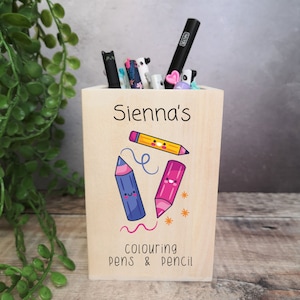 May include: A wooden pencil holder with the name "Sienna's" printed on the front. The holder features a cartoon illustration of three pencils with happy faces and the text "colouring pens & pencil".