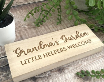 Könnte beinhalten: Rechteckiges Holzschild mit den Worten "Grandma's Garden" und "Little Helpers Welcome" in kursiver Schrift graviert. Das hellfarbene Schild liegt auf einer Holzoberfläche, mit Grünpflanzen und einem kleinen weißen Topf im Hintergrund.