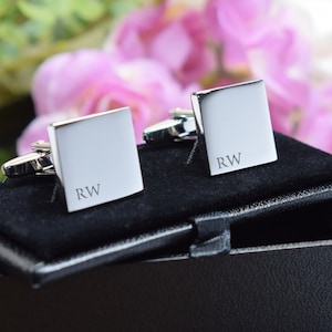 May include: Silver square cufflinks, each engraved with the initials "RW", displayed in a black presentation box with a velvet lining. The background shows pink and green flowers.
