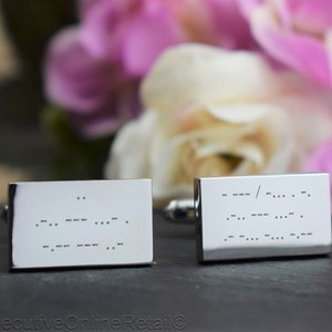 May include: A pair of rectangular silver cufflinks with Morse code engraving. The cufflinks are displayed against a backdrop of blurred pink and white flowers. The Morse code is etched into the polished metal surface.
