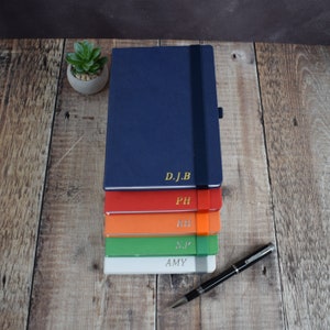 May include: A stack of personalized notebooks in various colors: navy blue, red, orange, green, and white. Each notebook has gold lettering with initials or a name. A black pen with silver accents rests on a wooden surface.