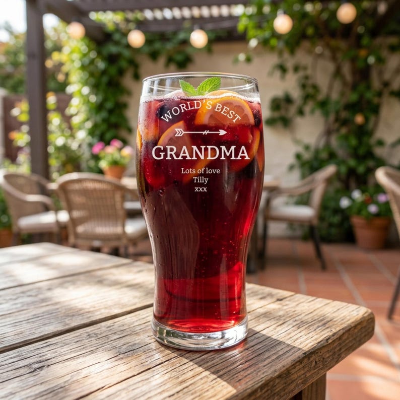 May include: A tall glass filled with a red beverage, garnished with orange slices and mint. The glass is etched with the words "WORLD'S BEST GRANDMA" and "Lots of love Tilly xxx". The glass sits on a wooden table outdoors.