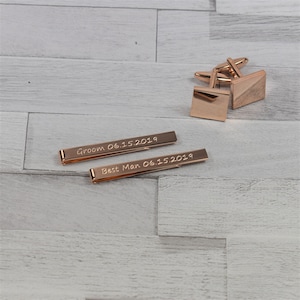 May include: Two rose gold tie bars with engraved text. The top tie bar reads "Groom 06.15.2019". The bottom tie bar reads "Best Man 06.15.2019".