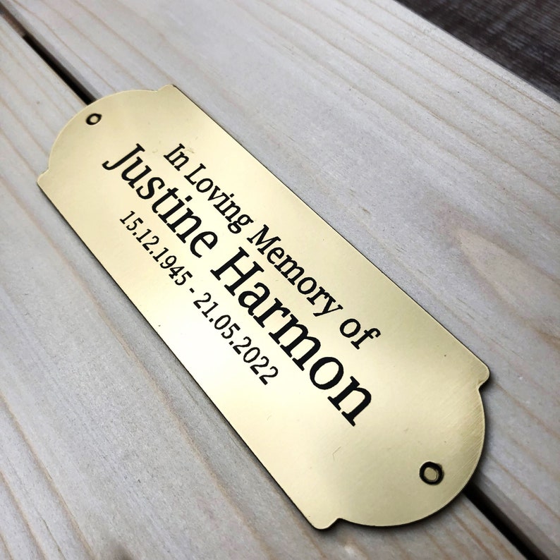 May include: A gold-colored metal plaque with rounded edges and a black inscription that reads "In Loving Memory of Justine Harmon 15.12.1945 - 21.05.2022".