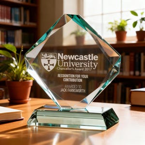 May include: A clear glass award with a diamond-shaped top and a rectangular base. The award features the Newcastle University logo and text that reads "Recognition for Your Contribution" and "Awarded to Jack Farnsworth."