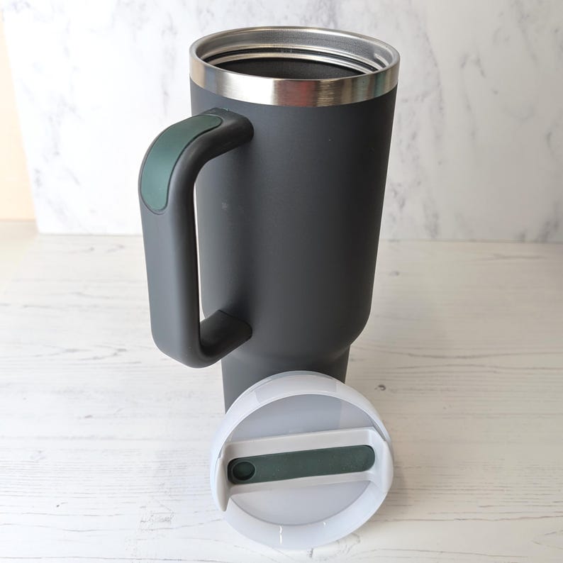 May include: A dark grey insulated travel mug with a handle and a stainless steel rim. The handle has a green accent. The white lid is off to the side, revealing a green slider. This mug is designed to keep drinks hot or cold.