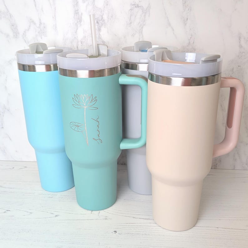 May include: Four insulated tumblers in pastel shades: light blue, mint green, pale pink, and off-white. Each tumbler has a handle, a silver rim, and a lid. The mint green tumbler features a floral design and the name "Sarah".