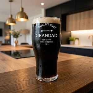 May include: A pint glass filled with dark beer and foam, with the words "WORLD'S BEST GRANDAD" in white lettering. Below the text are the words "Lots of love Millie and James xx". The glass sits on a wooden surface.