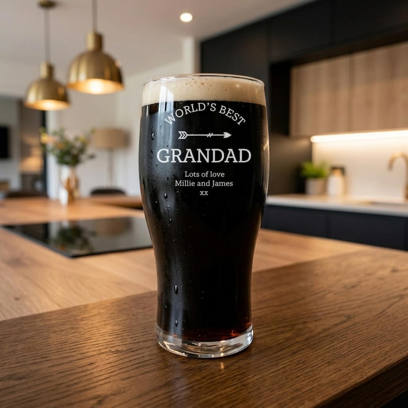 May include: A pint glass filled with dark beer and foam, with the words "WORLD'S BEST GRANDAD" in white lettering. Below the text are the words "Lots of love Millie and James xx". The glass sits on a wooden surface.