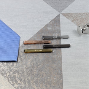 May include: Four personalised tie bars in different colours and finishes. The tie bars are engraved with the names "Andrew", "C.J.P", "CTP", and "25.11.2019".