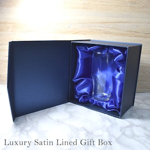 May include: A clear glass pint glass is displayed inside a blue satin-lined gift box. The box is open and the glass is visible inside.