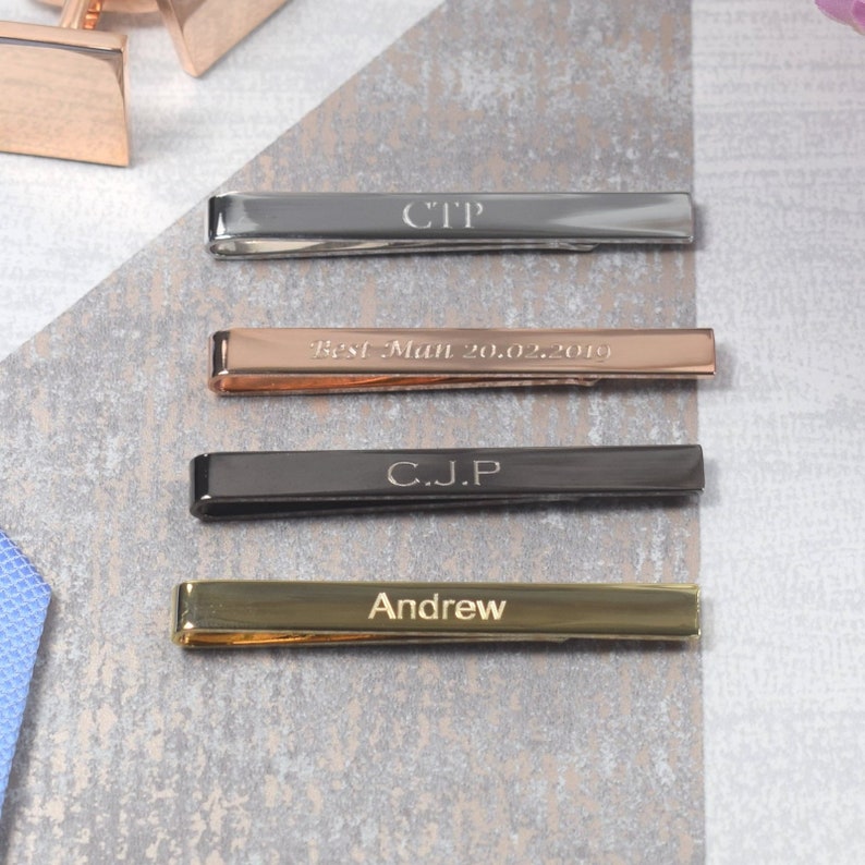 May include: Four tie bars in different colours and finishes. The top tie bar is silver with the initials "CTP" engraved on it. The second tie bar is rose gold with the text "Best Man 20.02.2019" engraved on it. The third tie bar is black with the initials "C.J.P" engraved on it. The bottom tie bar is gold with the name "Andrew" engraved on it.
