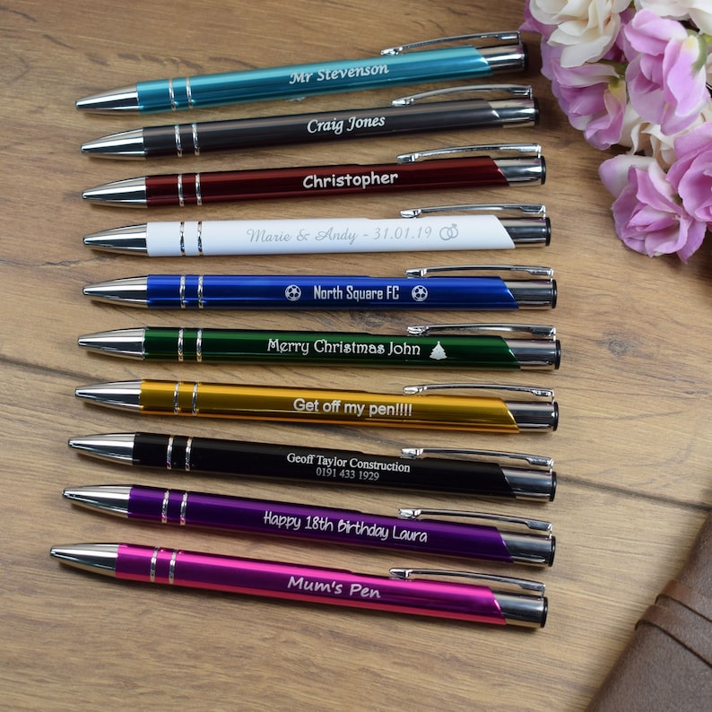 Puede incluir: Un conjunto de diez bol&iacute;grafos personalizados de varios colores con diferentes grabados. Los bol&iacute;grafos est&aacute;n dispuestos verticalmente sobre una superficie de madera. Los grabados incluyen nombres, fechas, mensajes y un logotipo de f&uacute;tbol.