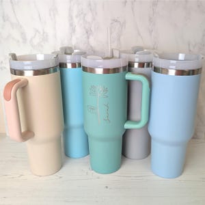 May include: An assortment of insulated tumblers in pastel shades such as cream, blue, and mint green. Each tumbler has a handle, a lid, and a silver rim. One tumbler is decorated with a floral design and the name "Sarah".