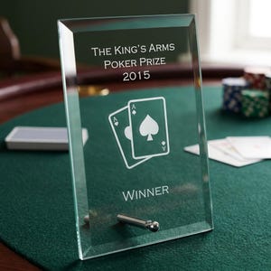 Puede incluir: Un premio de póquer de vidrio transparente con el texto "THE KING'S ARMS POKER PRIZE 2015" y un gráfico de dos cartas de juego. También se muestra la palabra "WINNER". El premio está sobre una superficie de fieltro verde.