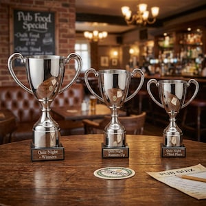 May include: Three silver trophies with handles and wooden bases sit on a wooden table. The trophies are labeled "Quiz Night - Winners", "Runners Up", and "Third Place". A coaster and a pub quiz sheet are also on the table.