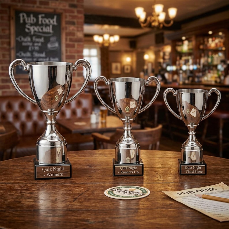 May include: Three silver trophies with handles and wooden bases sit on a wooden table. The trophies are labeled "Quiz Night - Winners", "Runners Up", and "Third Place". A coaster and a pub quiz sheet are also on the table.