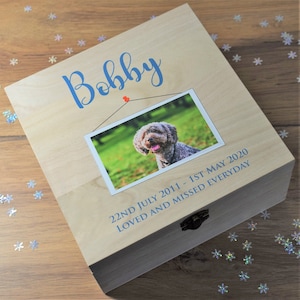 May include: A wooden box with a photograph of a dog, with the name "Bobby" in blue script. The photo is of a smiling dog. The text "22nd July 2011 - 1st May 2020 Loved and Missed Everyday" is printed below the photo in blue.