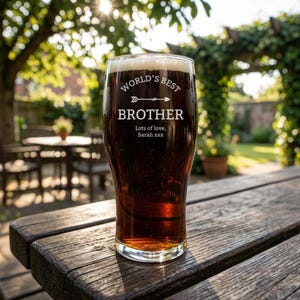 May include: A pint glass filled with dark beer, resting on a wooden table. The glass is etched with the words "WORLD'S BEST BROTHER" and a personalized message. The background features a sunny outdoor setting with greenery.