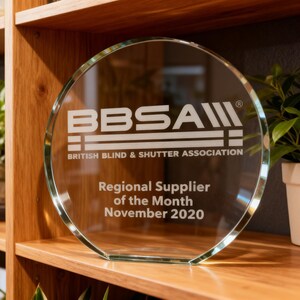 Może przedstawiać: Okrągła szklana nagroda z wygrawerowanym białym logo BBSA British Blind & Shutter Association. Poniżej logo znajduje się napis Regional Supplier of the Month November 2020. Nagroda posiada płaską podstawę zapewniającą stabilność.