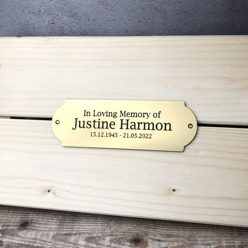 May include: Gold-colored metal plaque with the text "In Loving Memory of Justine Harmon 15.12.1945 - 21.05.2022".