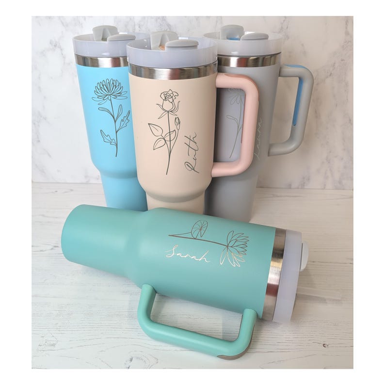 May include: Several insulated tumblers in pastel shades of blue, beige, and grey. Each tumbler is decorated with a floral illustration and a name in script. The tumblers have handles and lids, suitable for hot or cold drinks.