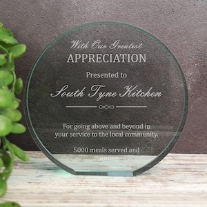 May include: A round, clear glass plaque with a white inscription. The inscription reads "With Our Greatest Appreciation Presented to South Tyne Kitchen For going above and beyond in your service to the local community. 5000 meals served and counting."