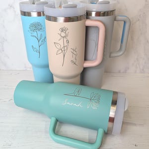 May include: Several insulated travel mugs in shades of blue, beige, and grey. Each mug has a floral design and a name. The mugs are equipped with handles and lids, ideal for hot or cold drinks.
