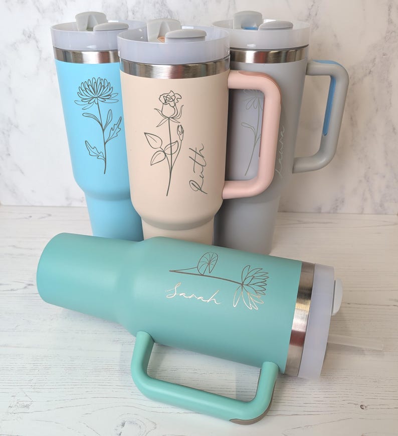 May include: Several insulated travel mugs in shades of blue, beige, and grey. Each mug has a floral design and a name. The mugs are equipped with handles and lids, ideal for hot or cold drinks.