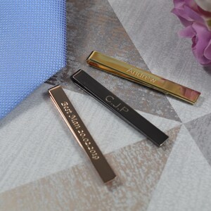 May include: Three tie clips with personalised engravings. The tie clips are gold, black and rose gold. The gold tie clip says "Andrew", the black tie clip says "C.J.P", and the rose gold tie clip says "Best Man 2022-2029".