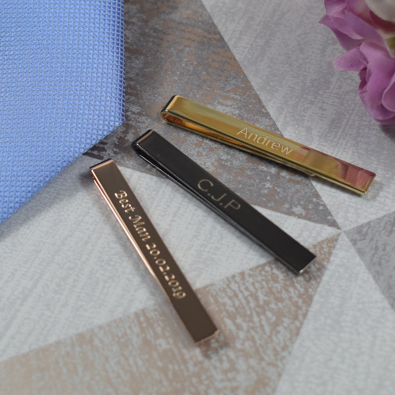 May include: Three tie clips with personalised engravings. The tie clips are gold, black and rose gold. The gold tie clip says "Andrew", the black tie clip says "C.J.P", and the rose gold tie clip says "Best Man 2022-2029".